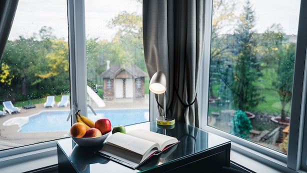 A view of a swimming pool through a window, a bowl of fruit, and an open book on a table.