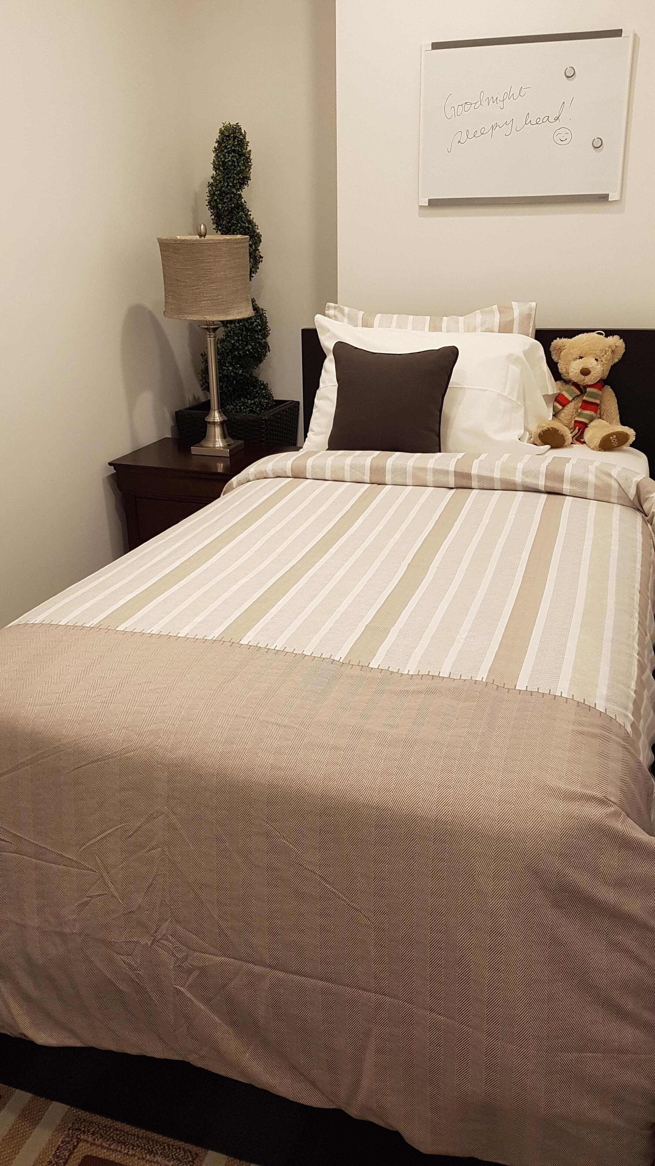 A neatly made bed with striped bedding, a bedside lamp, a small potted plant, and a teddy bear on the headboard.