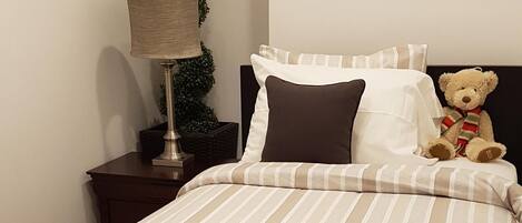 A neatly made bed with striped bedding, a bedside lamp, a small potted plant, and a teddy bear on the headboard.