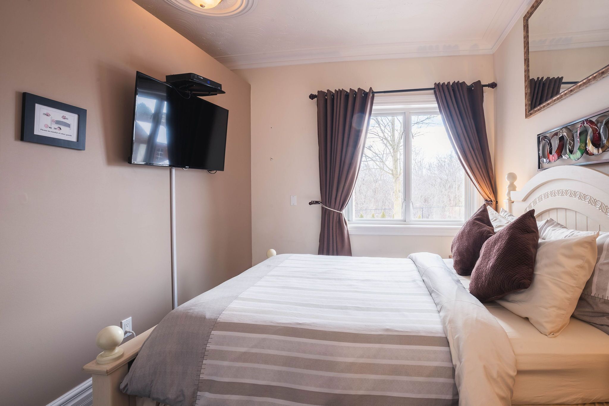 Un lit bien fait, recouvert d’une couverture à rayures, une table de chevet avec une lampe, un téléviseur à écran plat fixé au mur et une fenêtre avec des rideaux.
