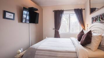 Un lit bien fait, recouvert d’une couverture à rayures, une table de chevet avec une lampe, un téléviseur à écran plat fixé au mur et une fenêtre avec des rideaux.