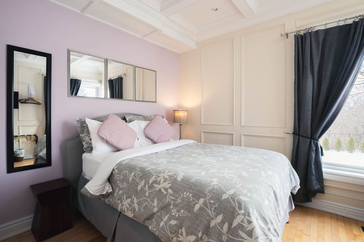 Un dormitorio con cama, dos espejos, una mesita de noche, una lámpara y una ventana con cortinas.