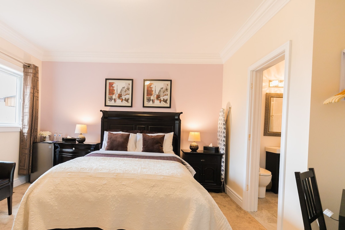 Quarto de hotel com uma cama grande identificada como 'Queen Bed', duas mesas de cabeceira com abajures, uma cadeira, uma escrivaninha e um espelho.