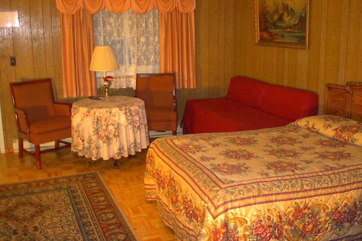 A room with a bed, two chairs, a small table with a lamp, and a painting on the wall.