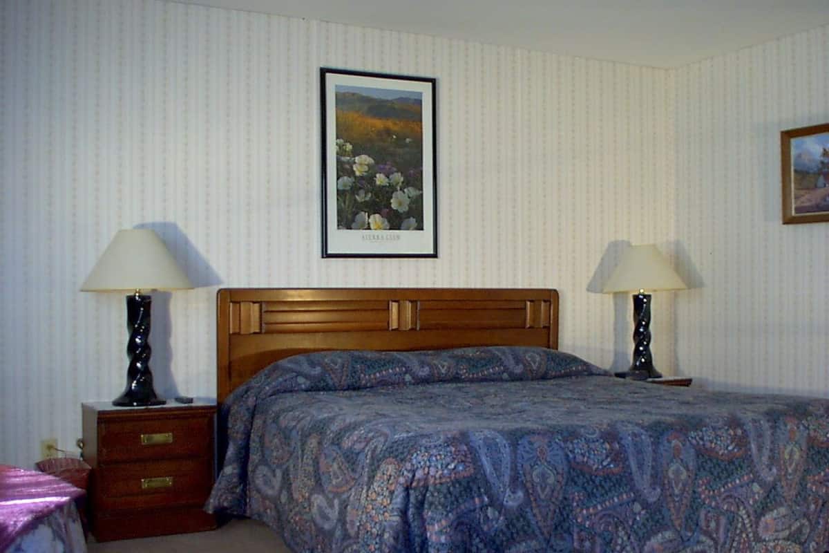 A bedroom with a wooden headboard, a patterned bedspread, two bedside tables with lamps, a framed picture on the wall, and a small drawer unit.