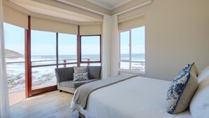 Een slaapkamer met een groot bed, een bank met kussens en uitzicht op het strand door grote ramen.