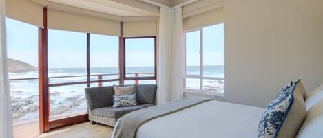 Un dormitorio con una cama grande, un sofá con cojines y vista a la playa a través de amplios ventanales.