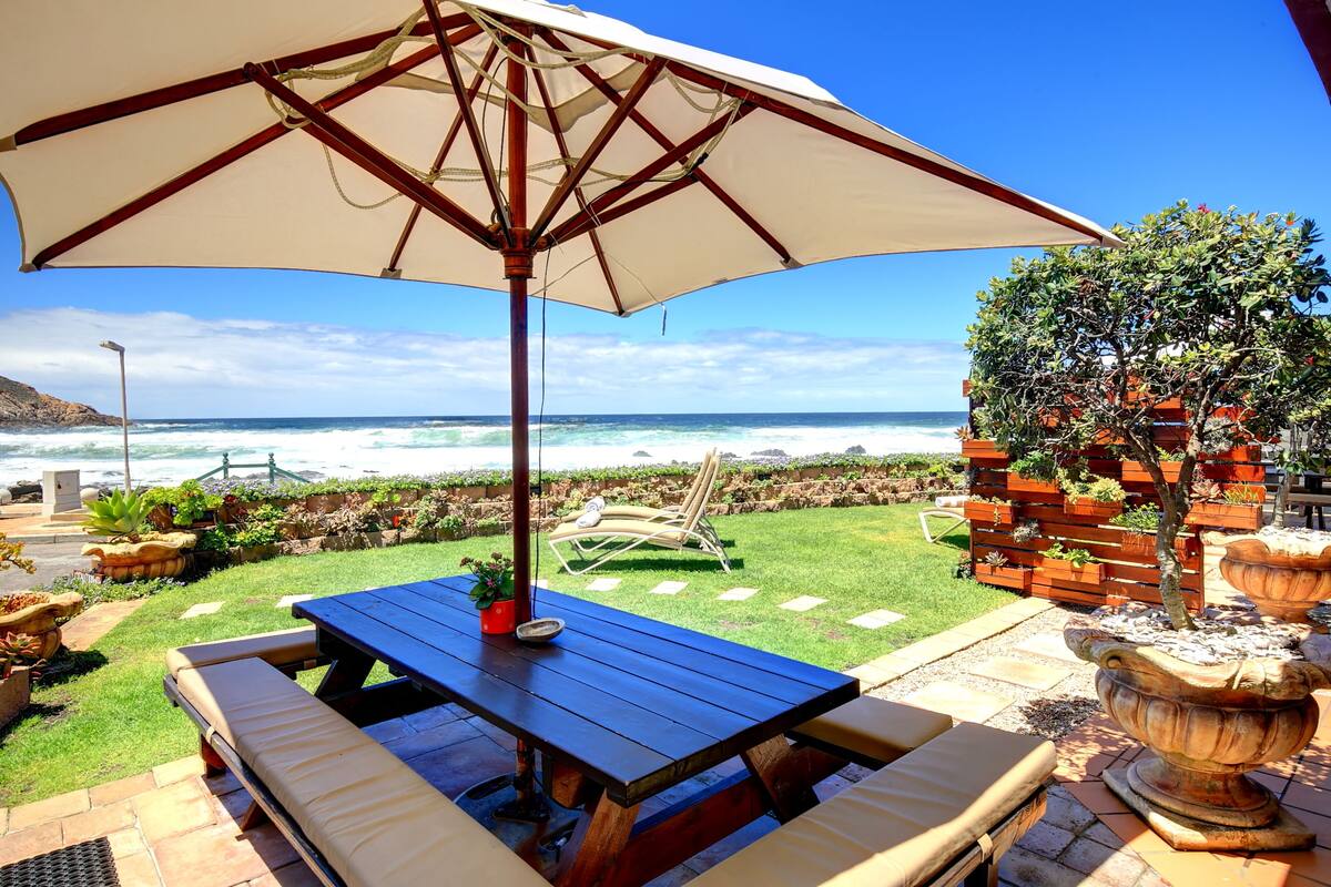 Een terras met tafel en stoelen, een grote parasol en uitzicht op zee.