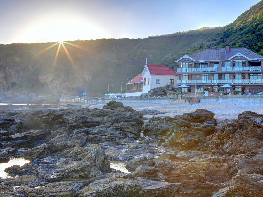 A coastal hotel with a red-roofed building, a balcony, and a view of the sea and hills.