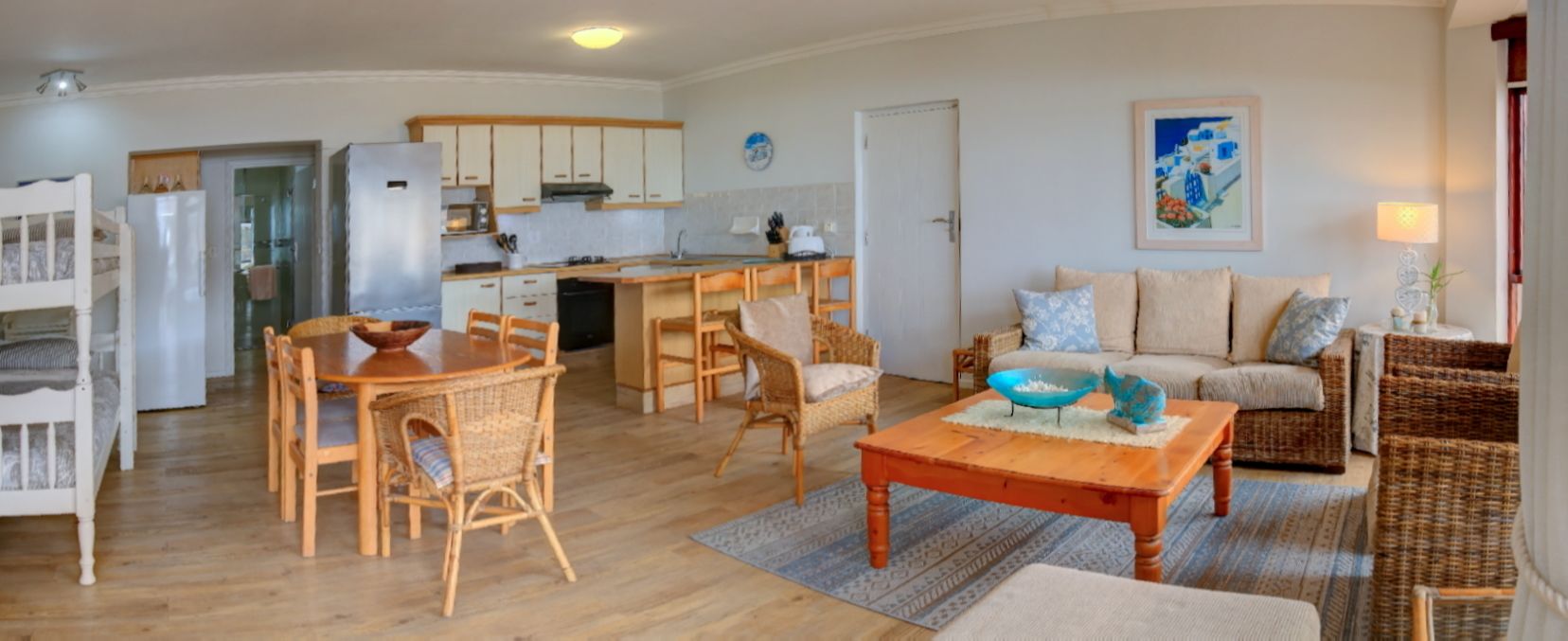 A spacious living area with a wooden dining table, wicker chairs, a sofa, and a kitchen with white cabinets and a refrigerator.