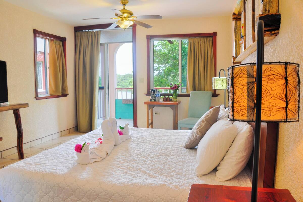 Habitación de hotel con cama, escritorio, silla, televisor y balcón con cortinas.