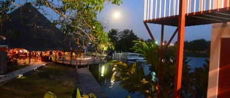 Un espacio de comedor junto al río con una terraza de madera, un puente y una montaña al fondo.