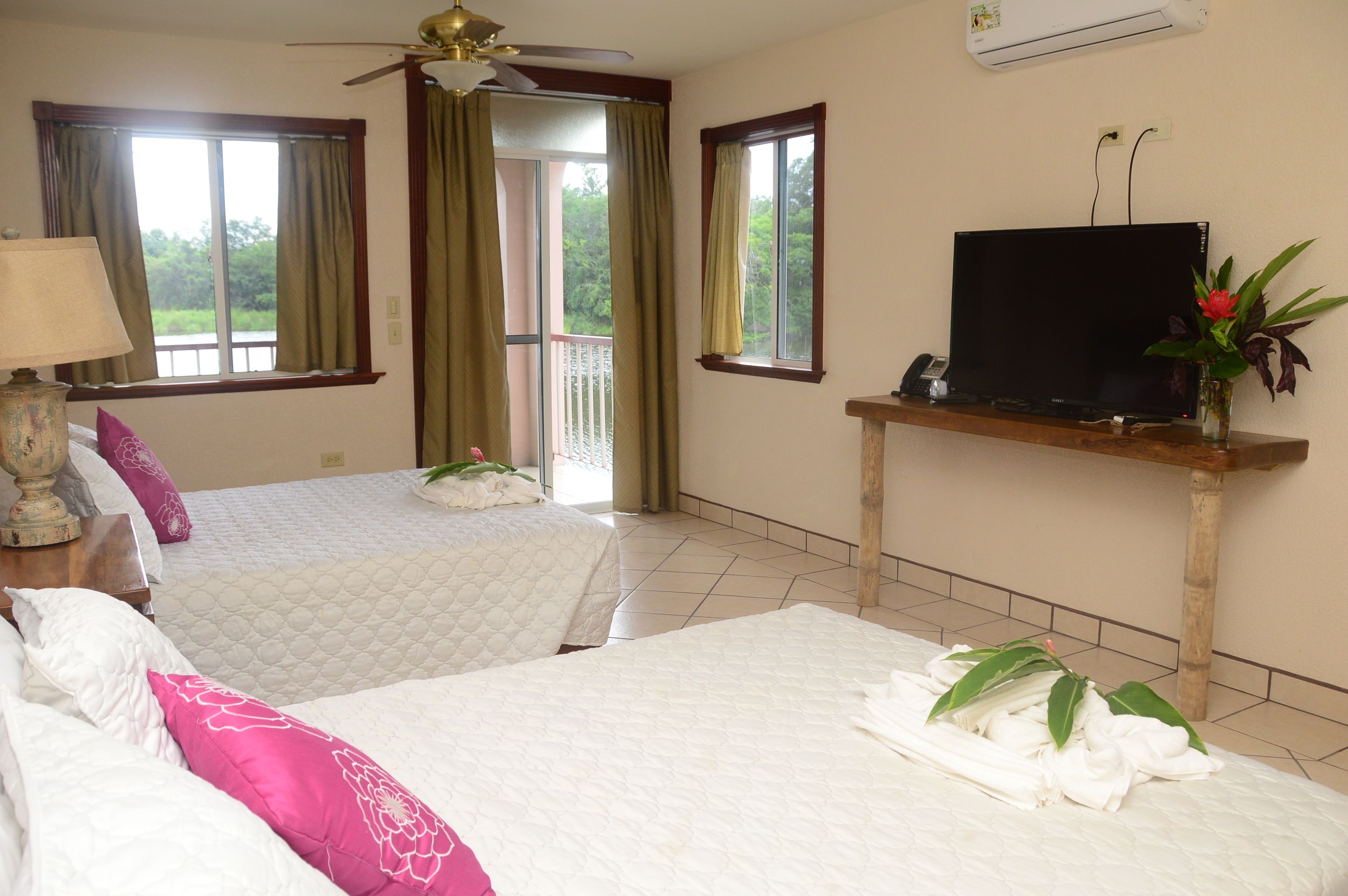 A hotel room with two beds, a TV on a wooden stand, and a balcony with curtains.