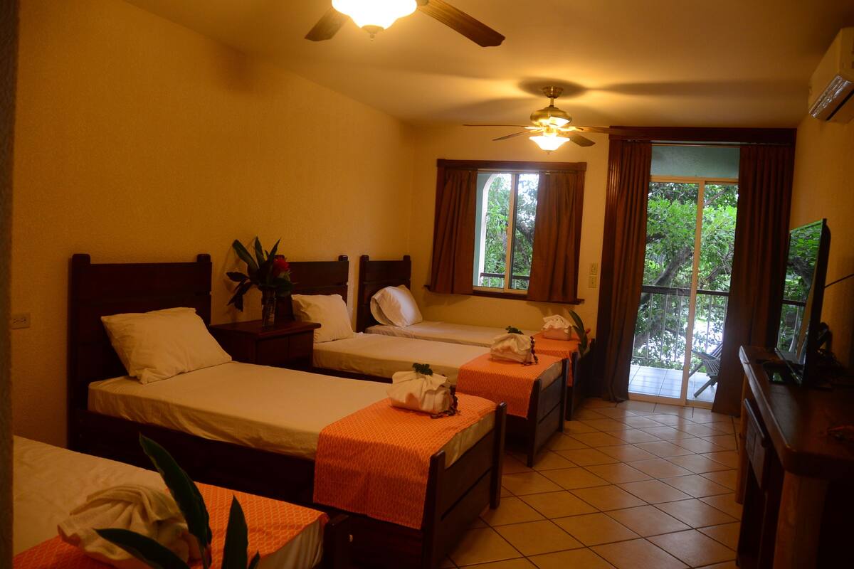 Habitación de hotel con tres camas individuales, ventilador de techo, televisión y ventana con vistas a zonas verdes.