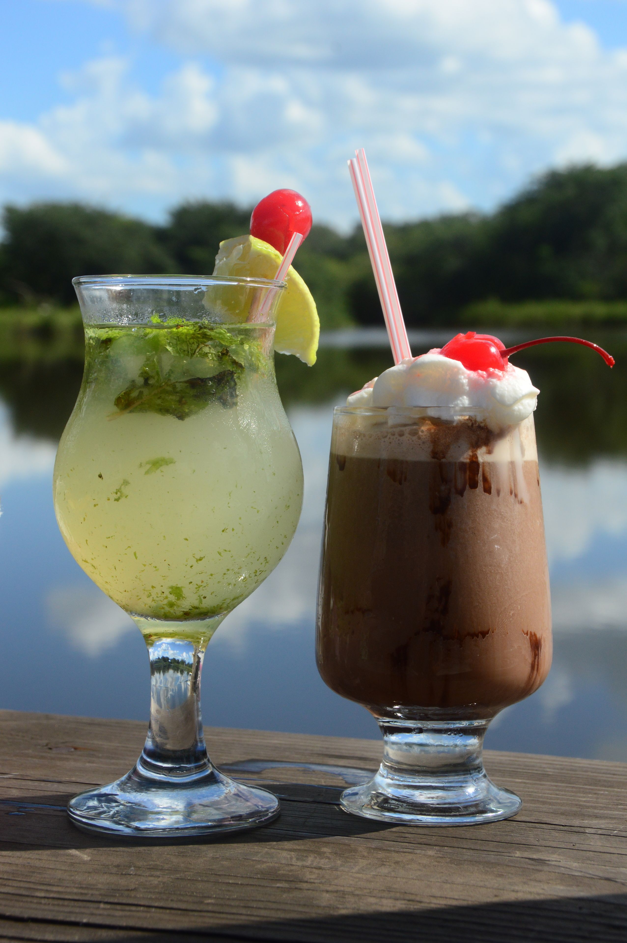 Two drinks on a wooden table with a lake and trees in the background.
