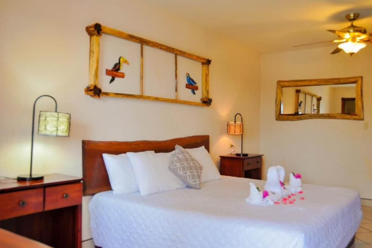 Une chambre d’hôtel comprenant un lit, une tête de lit en bois, une table de chevet avec une lampe, un miroir fixé au mur et des œuvres d’art.