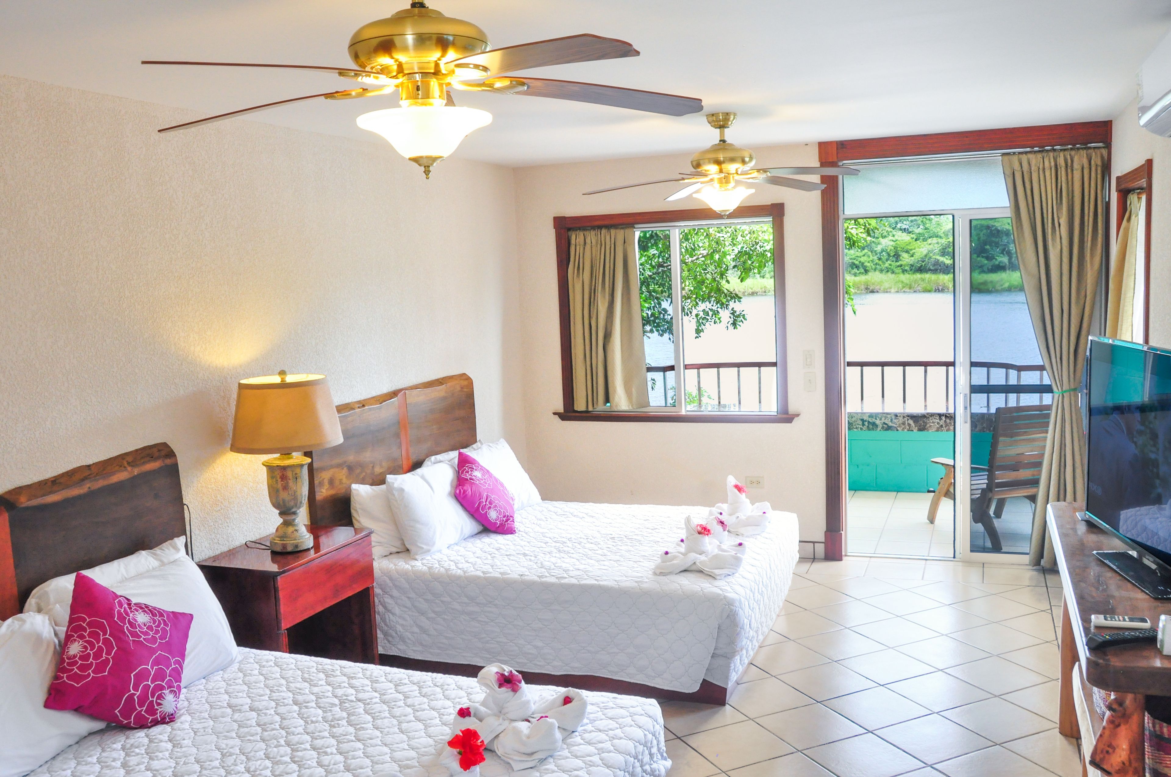 A hotel room with two beds, a ceiling fan, a TV, and a balcony with a view of greenery.