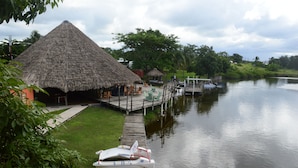 Eine reetgedeckte Hütte mit Außenplätzen, einem Anlegesteg und einem Boot sowie einem ruhigen Fluss.