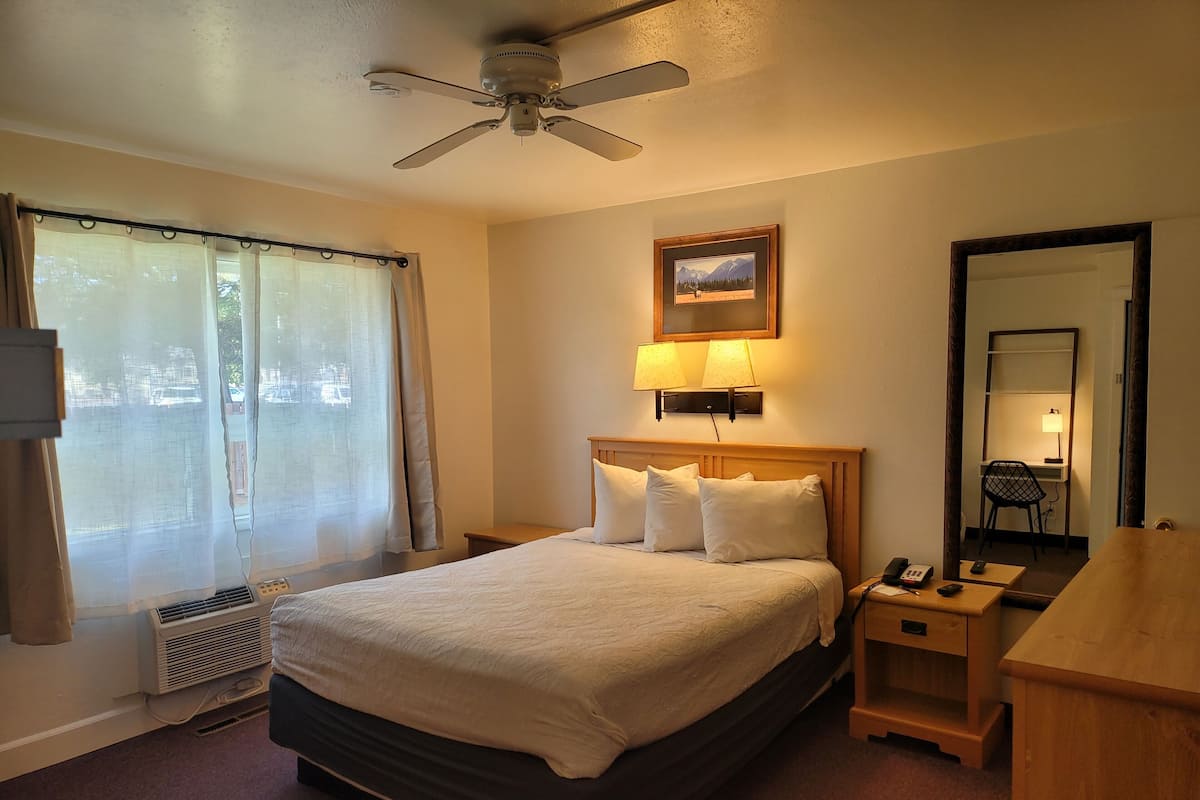 A hotel room with a bed, a desk, a chair, a ceiling fan, a window with curtains, a wall-mounted lamp, and a framed picture.