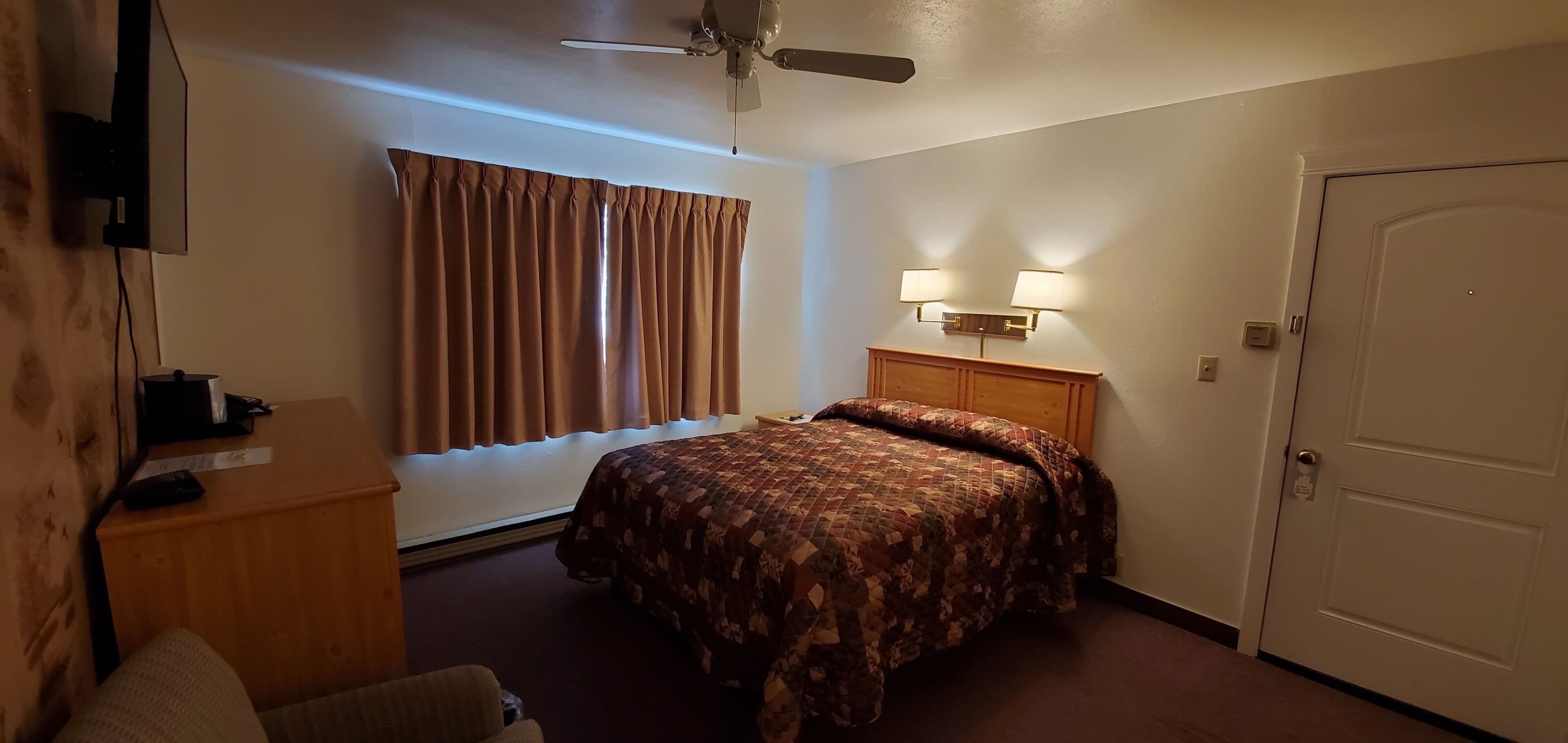 A hotel room with a bed, a desk, a chair, a ceiling fan, a window with curtains, and a door.