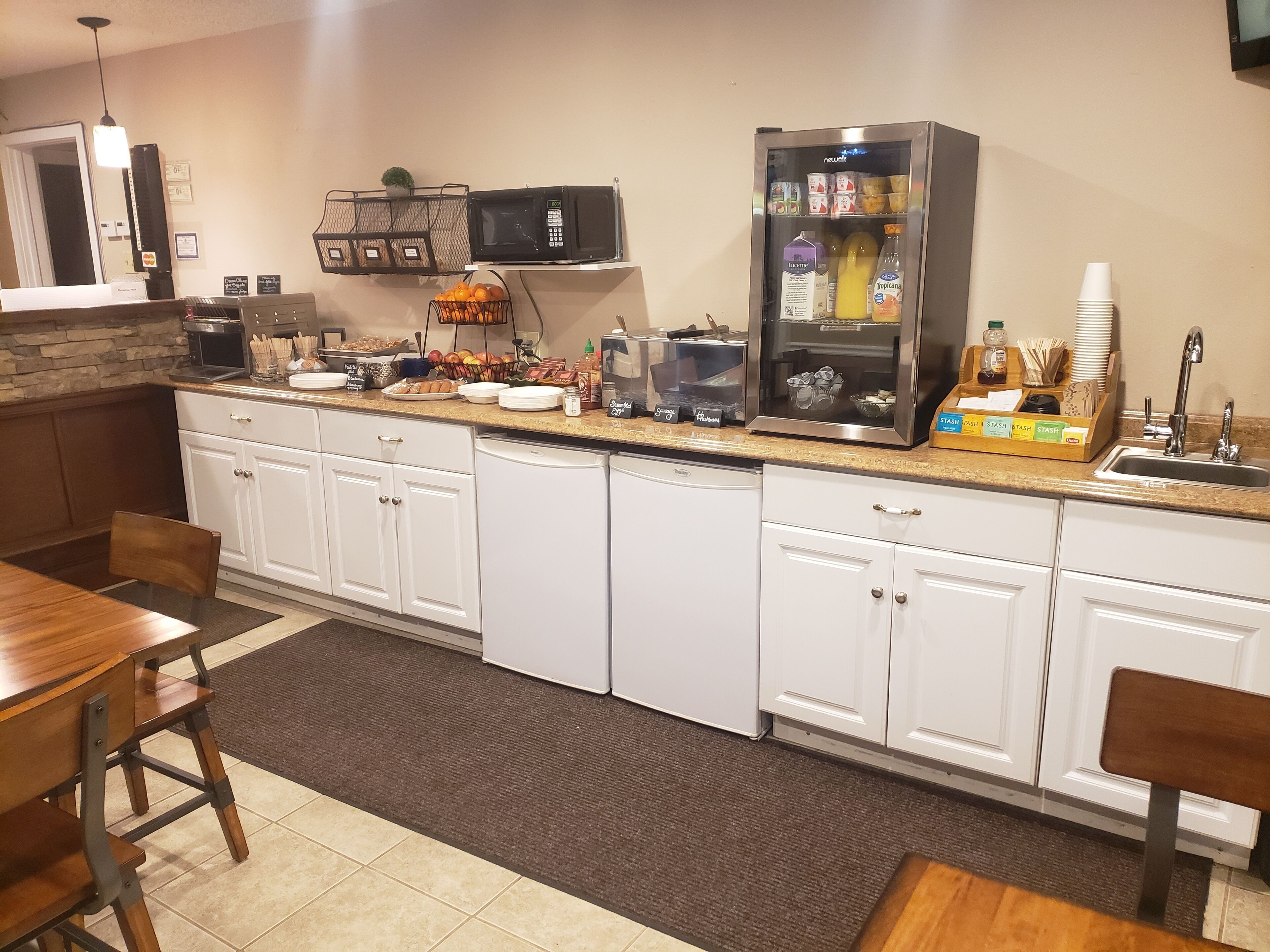 A kitchen with white cabinets, a microwave, a refrigerator, and a coffee machine. There are also fruits and beverages on the counter.
