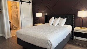 A modern bedroom with a large bed, two bedside tables with lamps, a dark headboard, and a sliding door leading to a shower area. - Bridge Street Inn (Baker City)