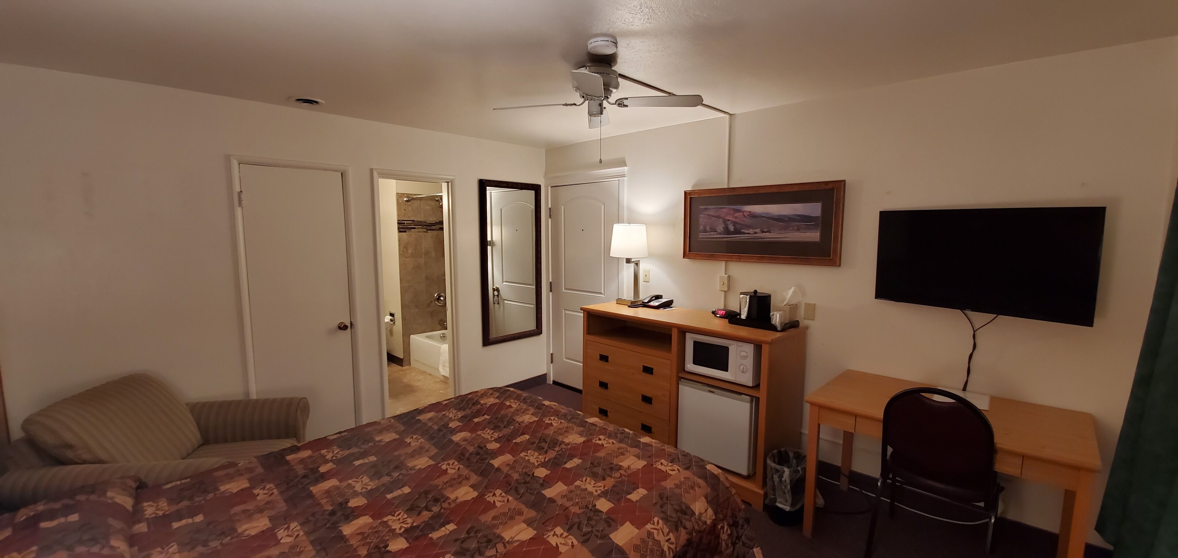 Quarto de hotel com cama, escrivaninha com computador, televisão, ventoinha no teto e banheiro visível por uma porta aberta.