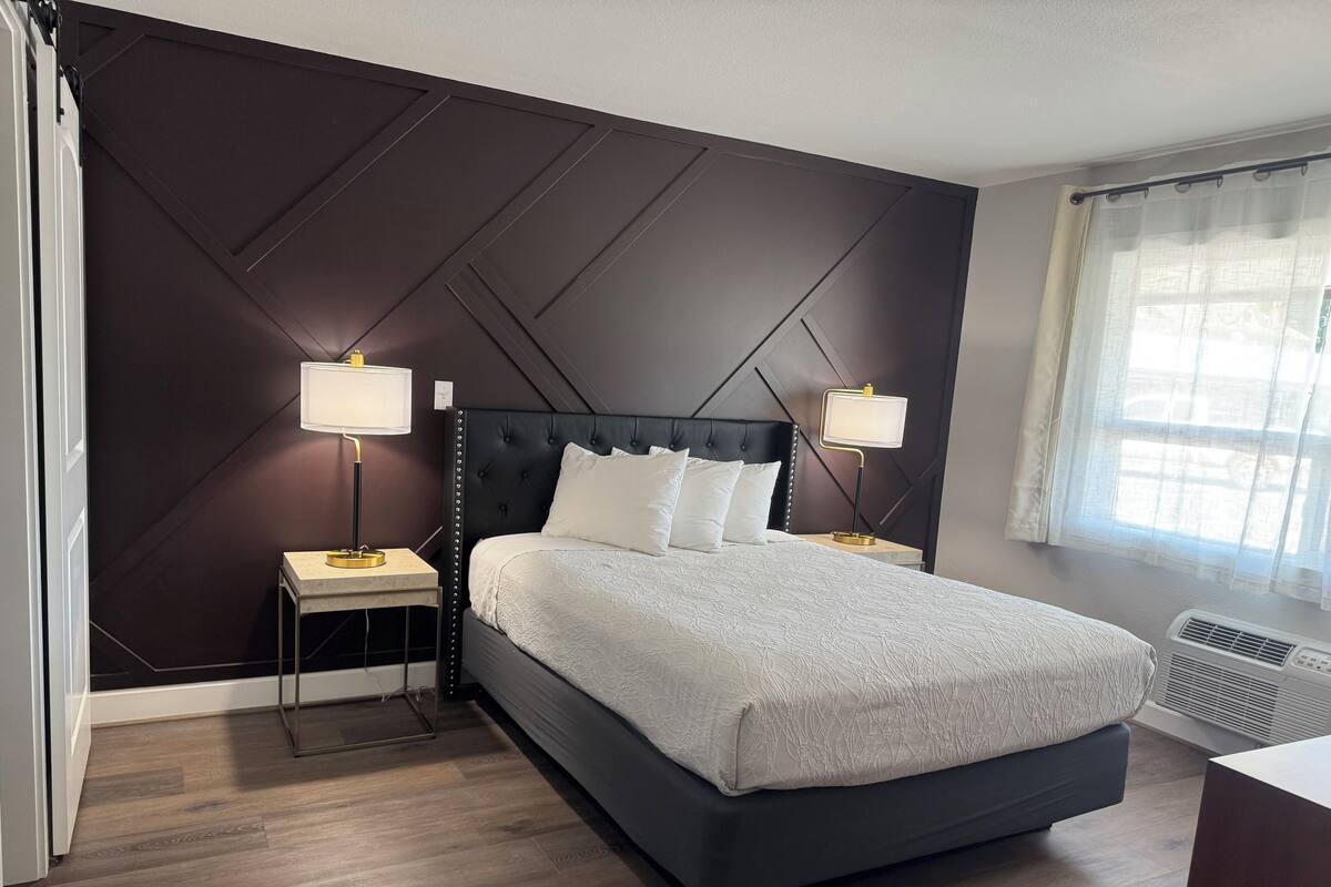 A modern hotel room with a dark wall, a large bed with white bedding, two bedside tables with lamps, and a window with sheer curtains.
