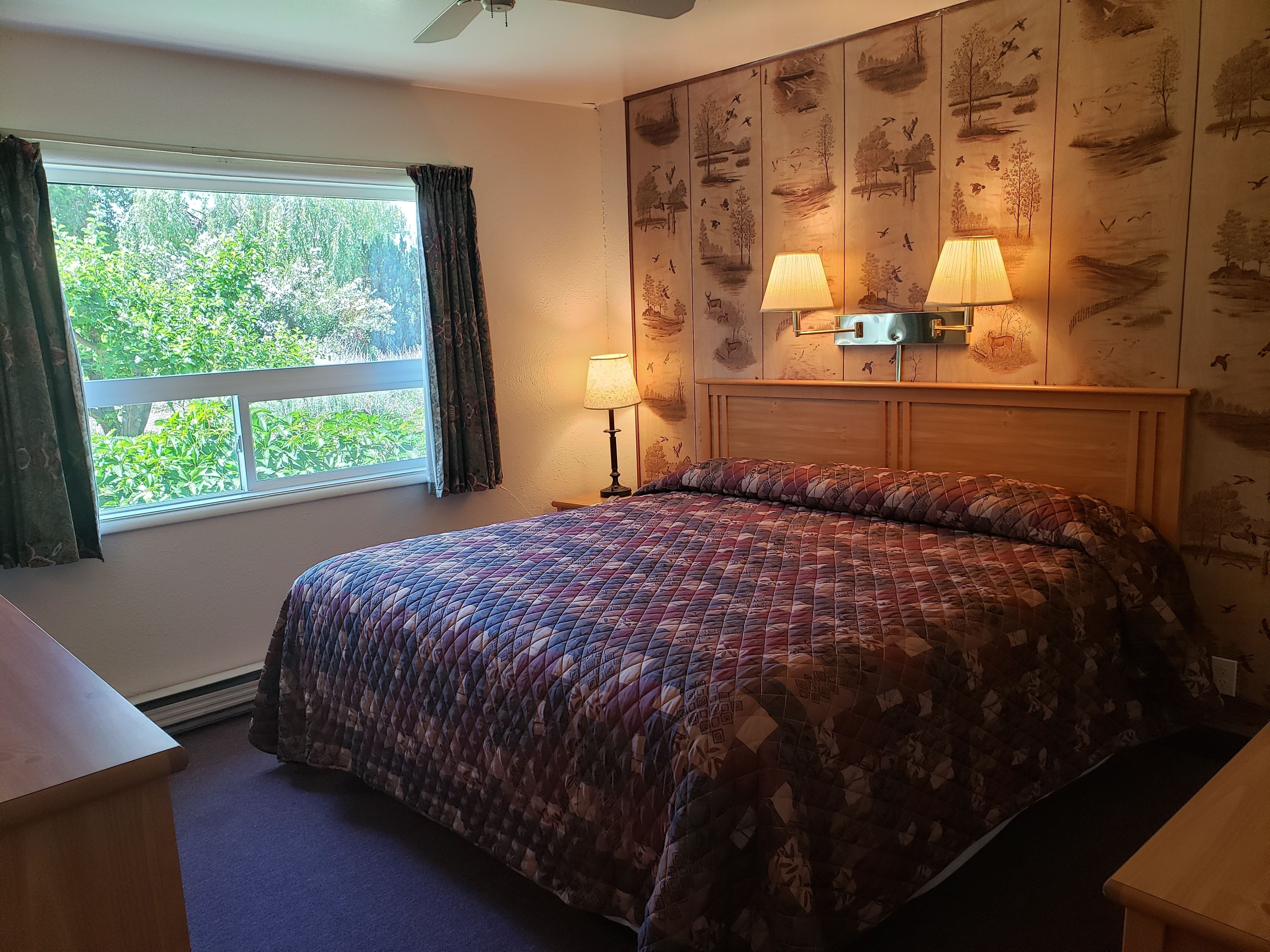 Un dormitorio con cama, ventana con cortinas, mesita de noche, dos lámparas de pared y un ventilador de techo.