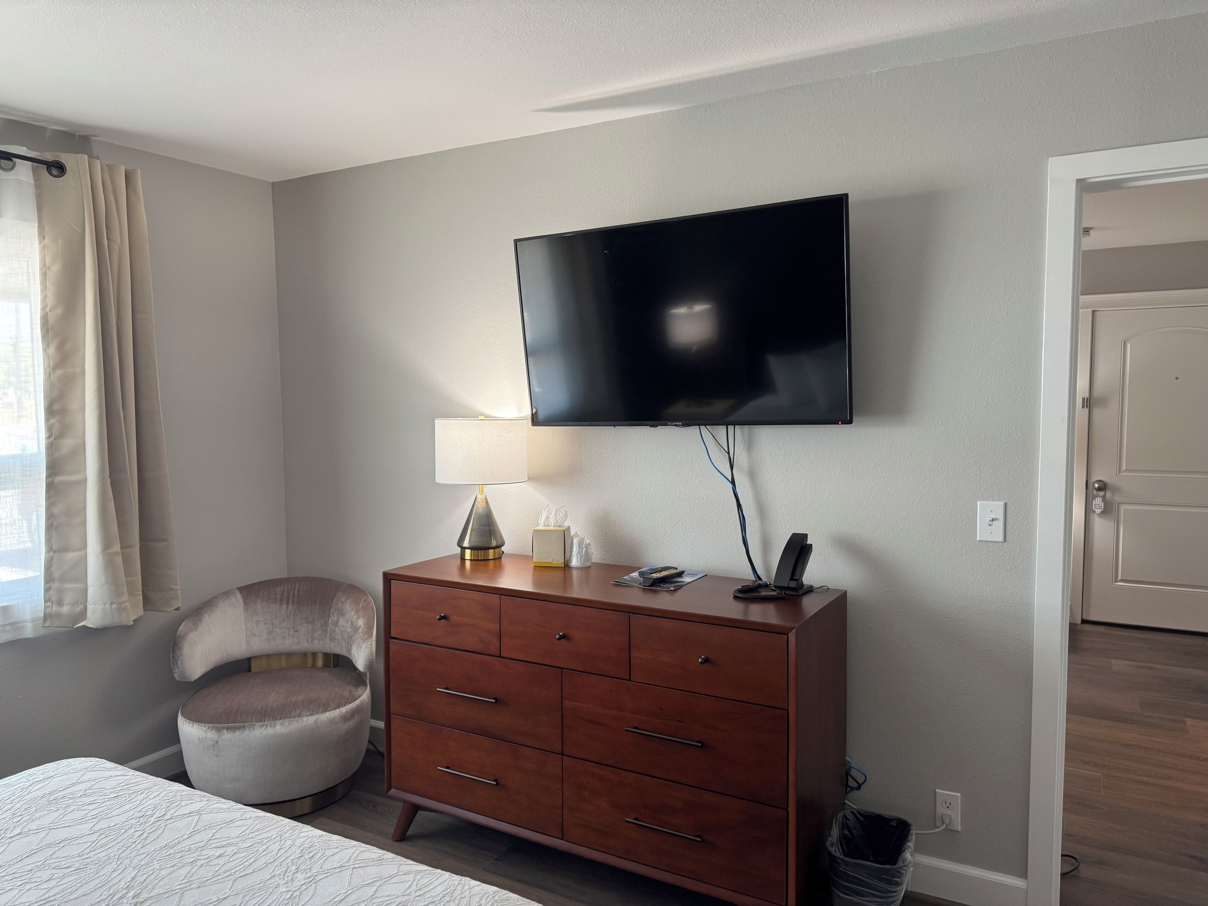 Een slaapkamer met een bed, een houten dressoir, een stoel, een aan de muur bevestigde televisie en een deur.
