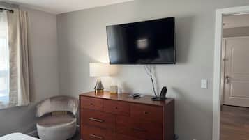 Een slaapkamer met een bed, een houten dressoir, een stoel, een aan de muur bevestigde televisie en een deur.