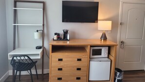 A compact hotel room with a wooden desk, a small refrigerator, a microwave, a TV, a lamp, a chair, and a trash bin. - Bridge Street Inn (Baker City)