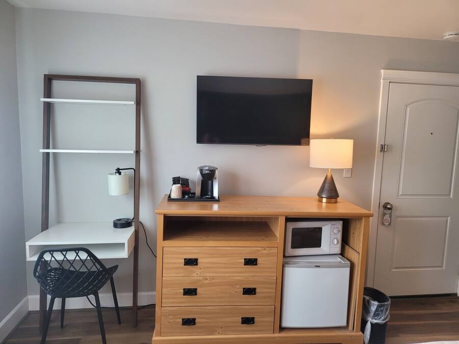A compact hotel room with a wooden desk, a small refrigerator, a microwave, a TV, a lamp, a chair, and a trash bin.