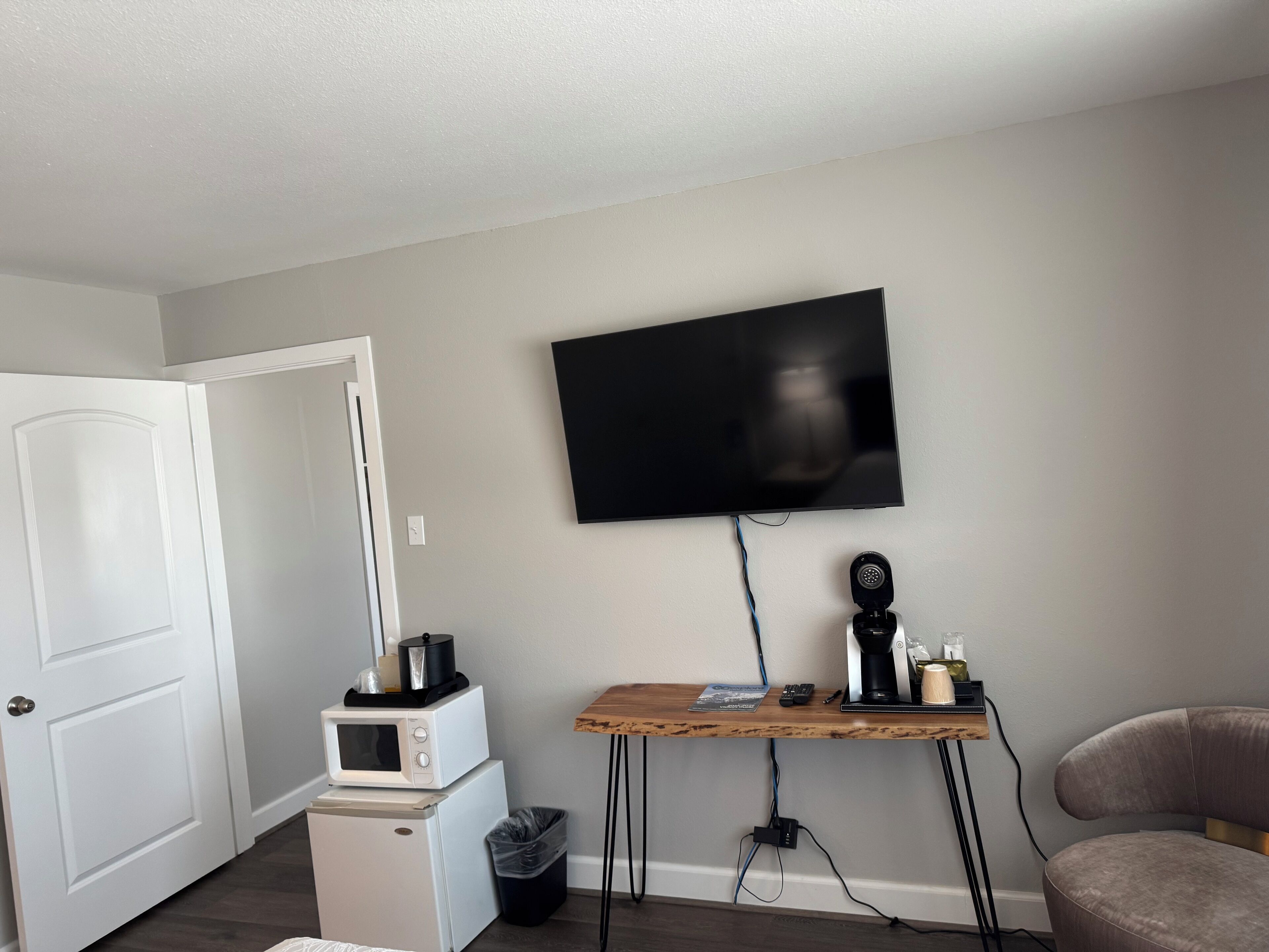 A compact living space with a flat-screen TV mounted on the wall, a small kitchenette with a microwave and mini-fridge, a wooden desk with a computer, and a cushioned chair.