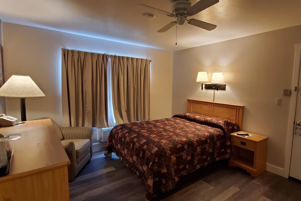 A hotel room with a bed, bedside lamps, a desk with a telephone, a chair, a ceiling fan, and a door.