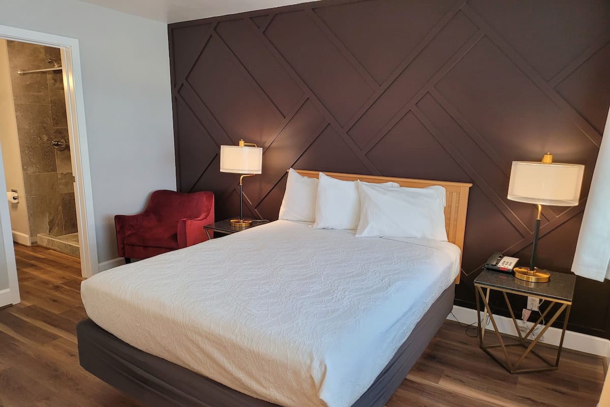 A hotel room with a large bed, two bedside lamps, a red chair, and a shower area.