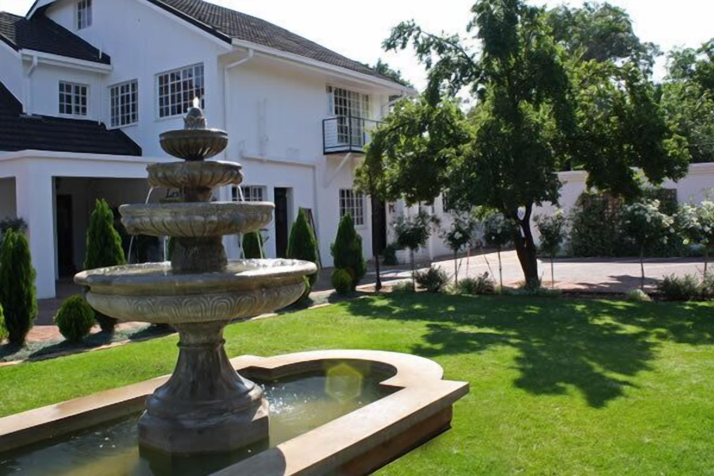 A white building with a multi-tiered fountain in the foreground and a tree to the right.