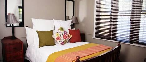 A bedroom with a bed, two bedside tables, a window with blinds, and a mirror on the wall.