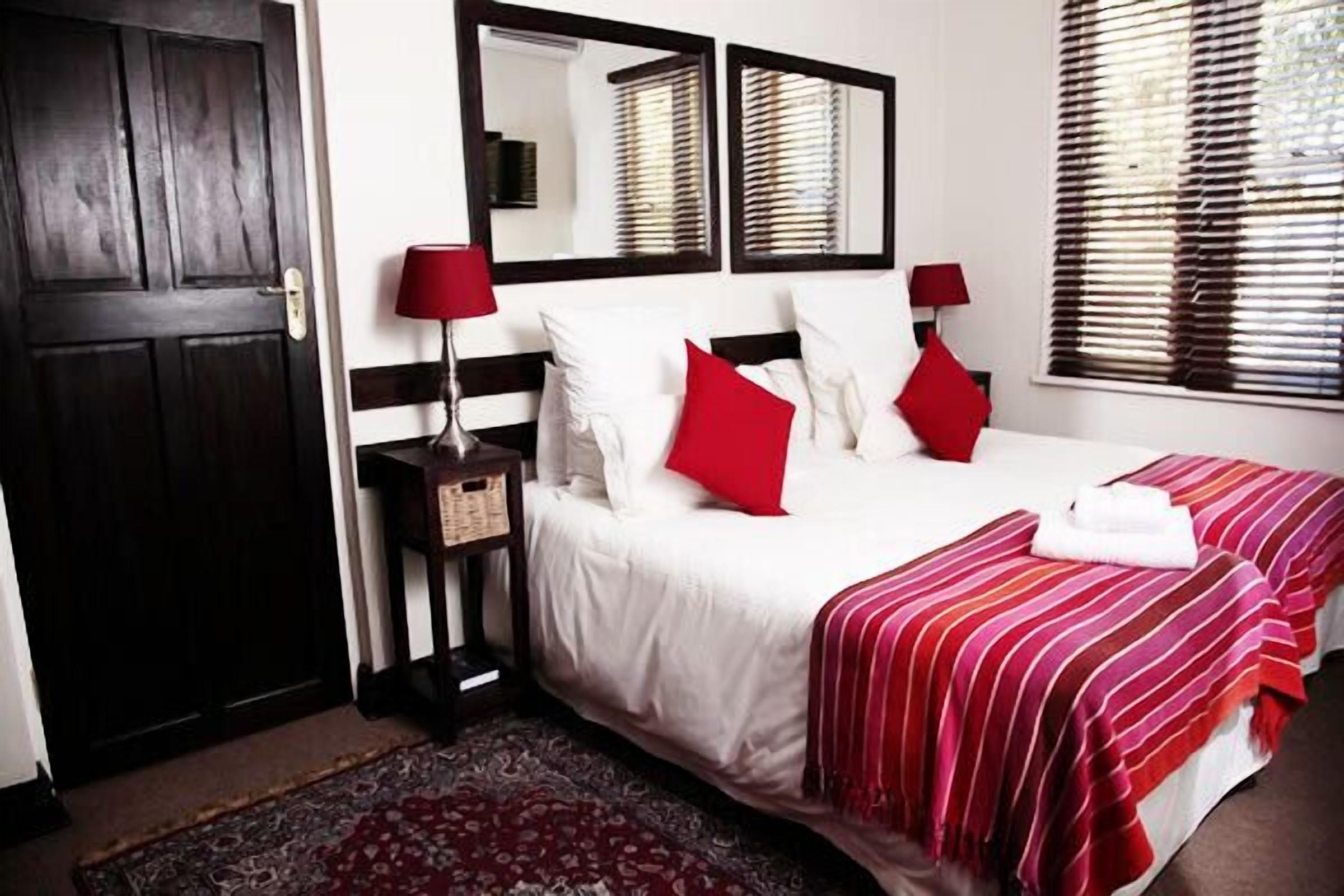 Une chambre d’hôtel avec une porte en bois sombre, un lit blanc avec des oreillers rouges, une couvre-lit rayée rouge et blanche, deux tables de chevet avec des lampes et un grand miroir au-dessus du lit.