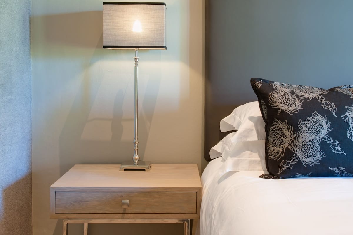 A neatly made bed with a white comforter and a dark patterned pillow, a bedside table with a lamp, and a wooden nightstand.