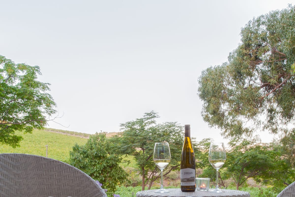 Wicker furniture, a bottle of wine, and glasses on a patio with a view of greenery.