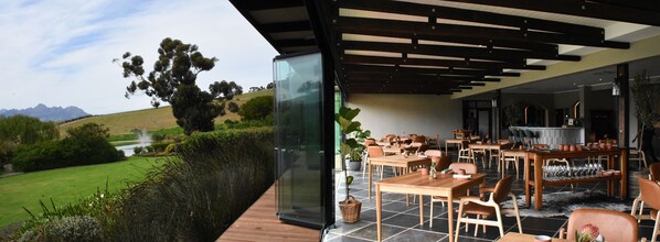 Ein Außenbereich mit Essbereich, Tischen, Stühlen und Blick auf einen Hügel und Bäume, teilweise durch eine Glasveranda geschützt.