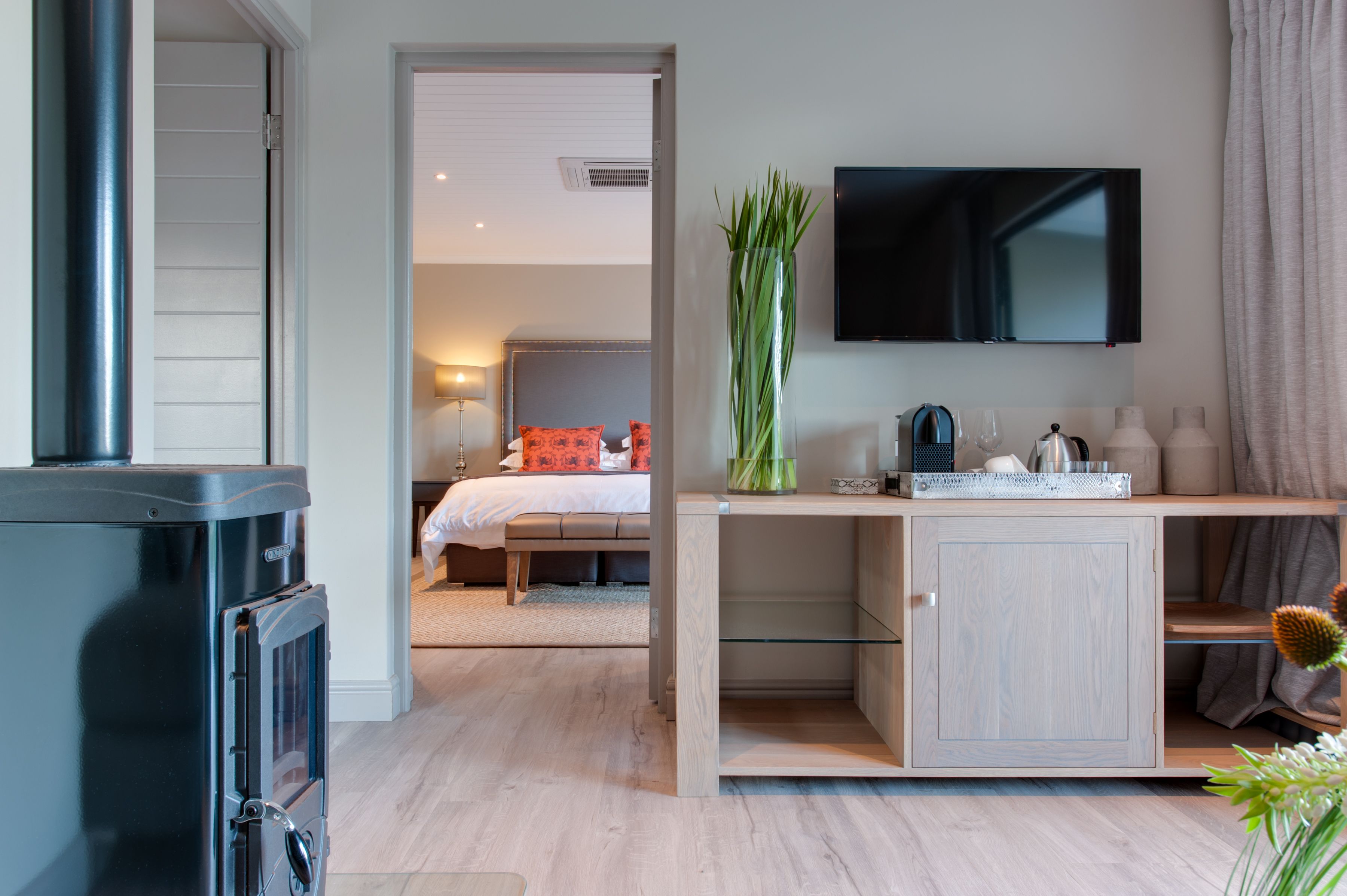 A modern hotel room with a wooden desk, a flat-screen TV, a bed with orange pillows, and a large vase of green plants.