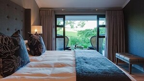 Quarto de hotel com uma cama grande, um banco, uma mesa e vista para áreas verdes lá fora.