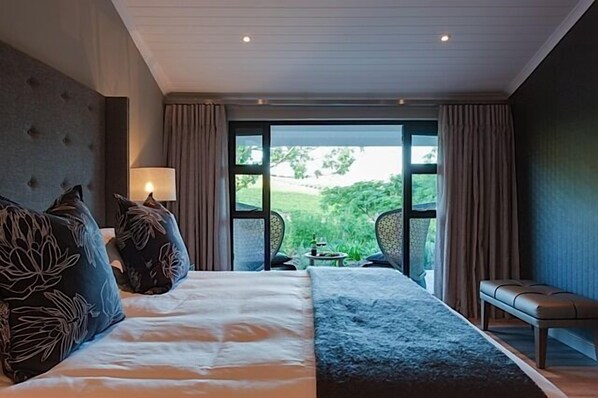 Une chambre d’hôtel avec un grand lit, un banc, une table et une vue sur la verdure environnante.