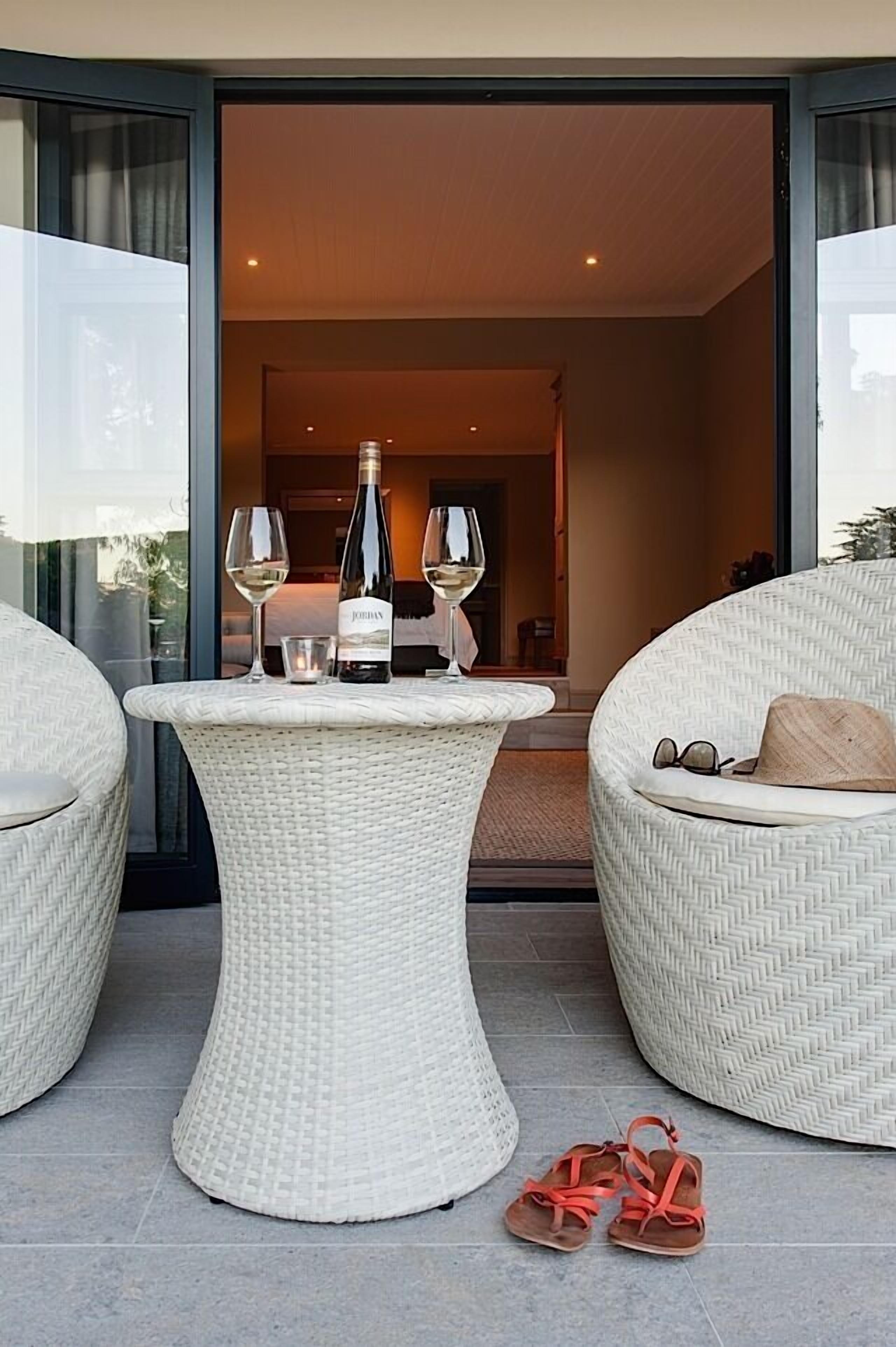 A wicker table with a bottle and glasses, a pair of sandals, and a wicker chair with a cushion.
