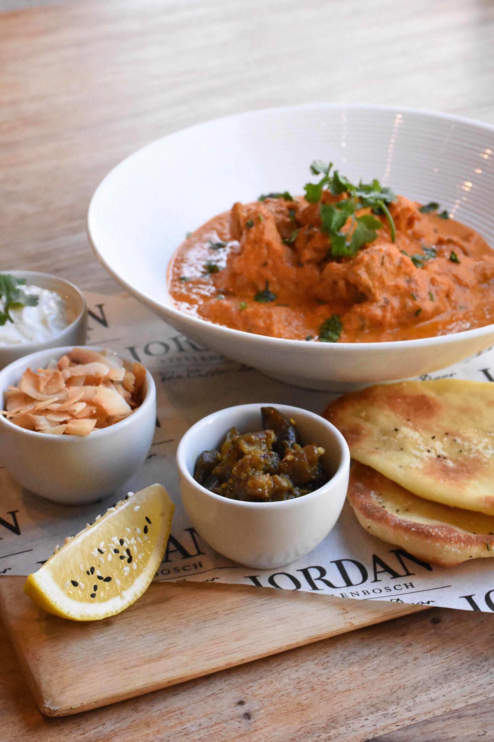 Una mesa con un tazón de curry, dos tazones pequeños de acompañamientos, un gajo de limón y pan plano.