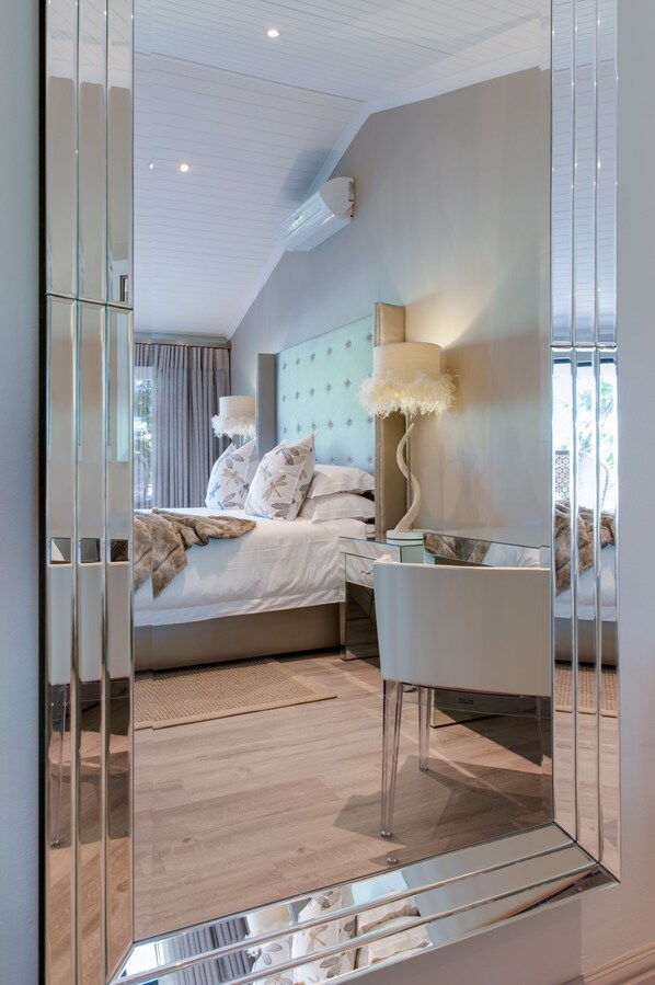 A modern bedroom with a large bed, a glass desk, and a mirror reflecting the room.