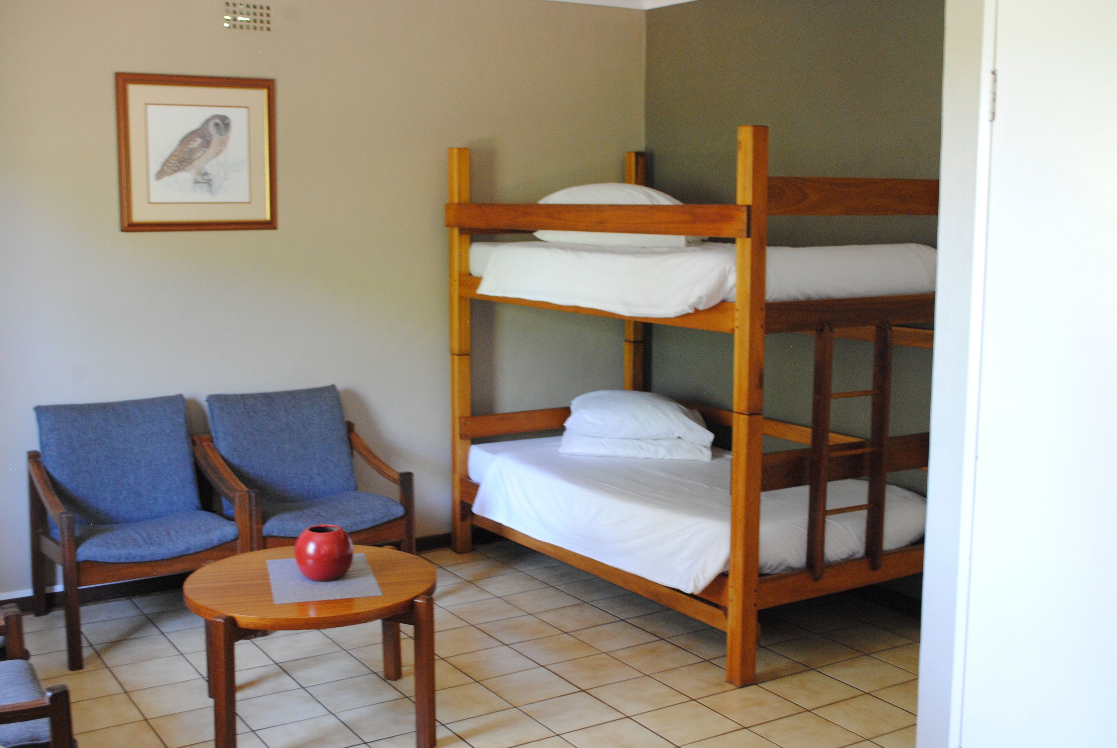 Una cama litera con ropa de cama blanca, dos sillones azules, una mesita de madera con un jarrón rojo, y un cuadro con un búho en la pared.
