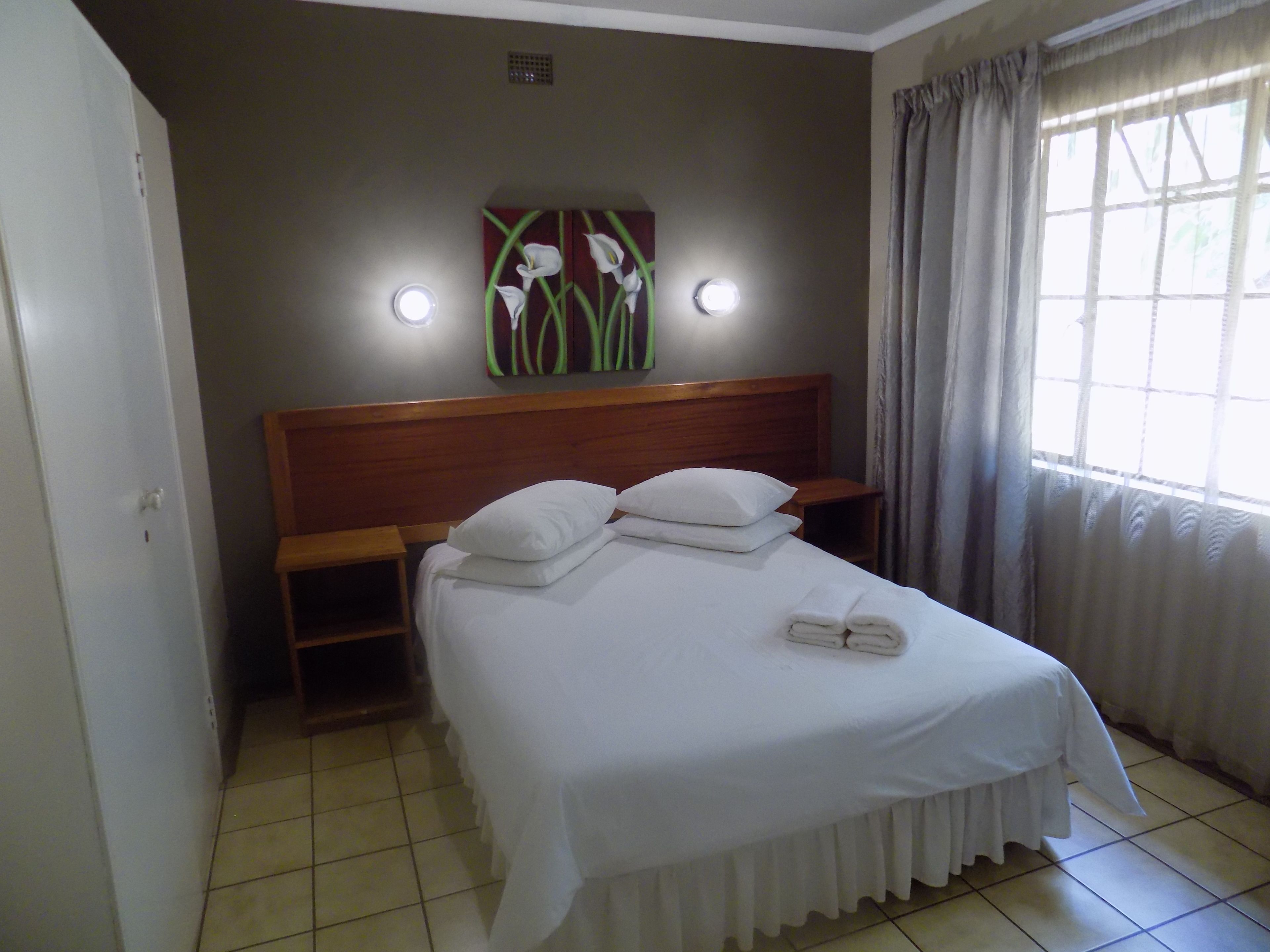 A neatly made bed with pillows and a folded towel, a wooden headboard, a small bedside table, and a wall-mounted light fixture.